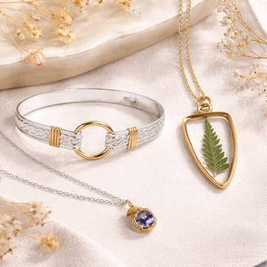 Artist crafted jewelry collection featuring a silver and gold bracelet, botanical fern pendant necklace, and gemstone necklace on a soft neutral background.