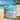 Jar of 'Beach Babe' whipped body scrub on a sandy beach with ocean and palm trees in the background.