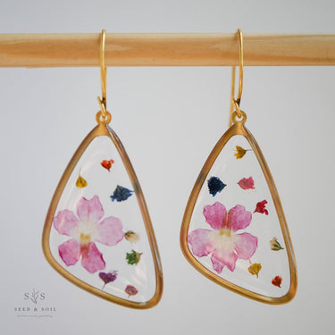 Gold-trimmed earrings with pressed flowers on a wooden rod against a light background