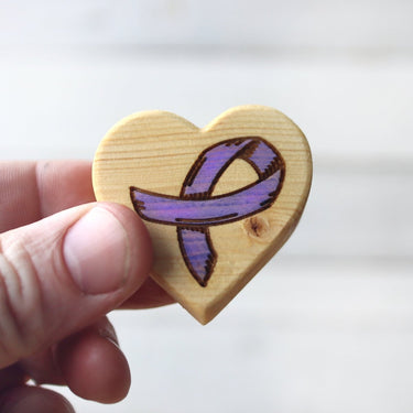 Wooden heart-shaped object with a purple ribbon design held by a hand against a light background