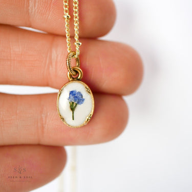 Gold necklace with a pendant featuring a blue flower held between fingers.