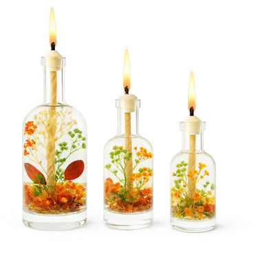 Three glass bottles with lit candles inside, containing floral and leaf decorations on a white background.