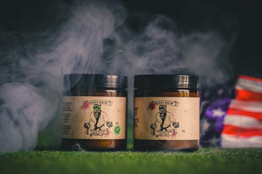 Two jars of 'Bentley Balm' on a grassy surface with smoke and an American flag in the background.