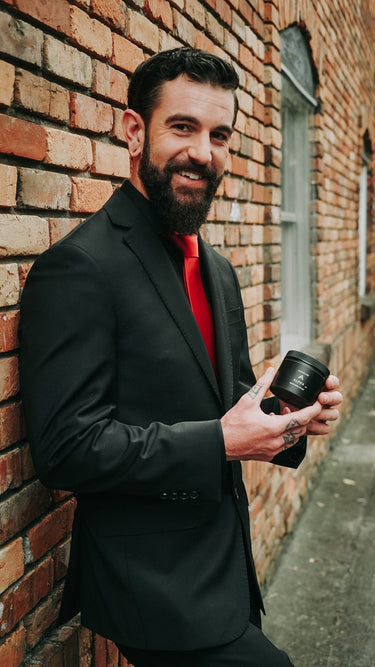 Man in a black suit with a red shirt holding a camera lens against a brick wall.