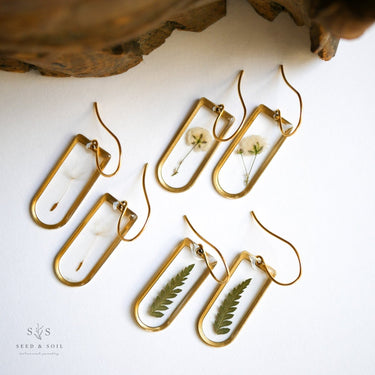 Gold-framed earrings with dried flowers and leaves on a white surface.