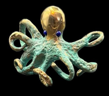 A small octopus Cuff with a metallic gold head and tentacles, covered in a textured turquoise-green patina, with two small dark blue bead eyes, set against a dark gray background.