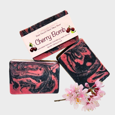 Three bars of cherry-flavored Natural Goat Milk Soap with a cherry bomb label, accompanied by pink flowers on a white background.