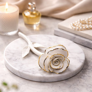 Decorative white rose with gold accents on a marble surface, surrounded by a candle and jewelry.