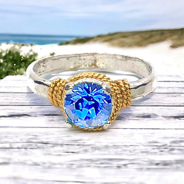 Ring with blue gemstone and gold accents on a wooden surface with a blurred natural background