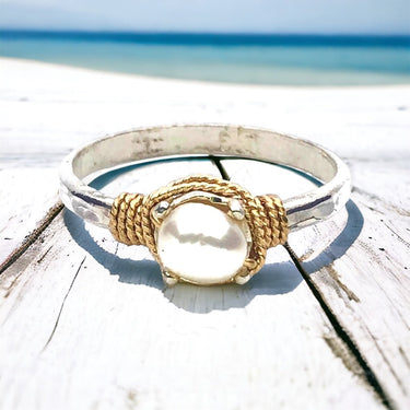 Silver ring with a gold and pearl design on a wooden surface with a beach background