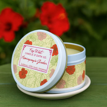 Candle in a tin with floral design and visible brand name on a green surface with red flowers in the background