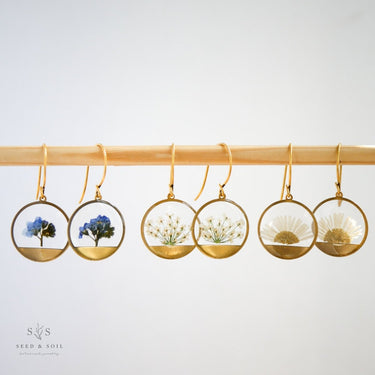 Set of six gold earrings with clear displays featuring different plants on a white background.