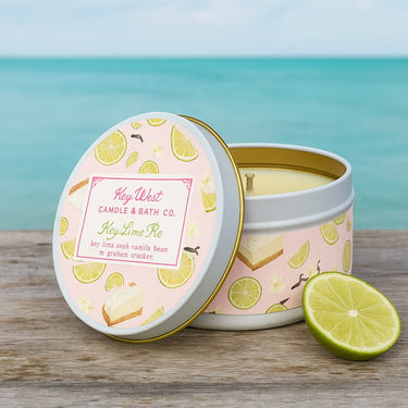 Candle in a decorative tin with lemon design, labeled 'Key West Candle & Bath Co.', on a wooden surface with a blue ocean background.