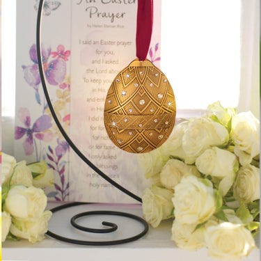 Decorative gold Easter egg ornament on a stand with flowers and a prayer card in the background.