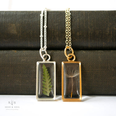 Two necklaces with pendants featuring pressed fern and dandelion seeds against a dark fabric background.