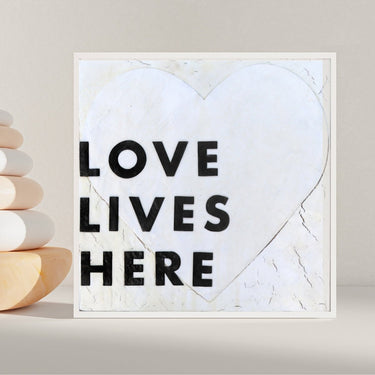 Framed heart-shaped sign with 'LOVE LIVES HERE' text on a neutral background