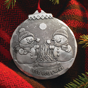 Silver Christmas ornament with two snowmen by a campfire on a red plaid fabric background.