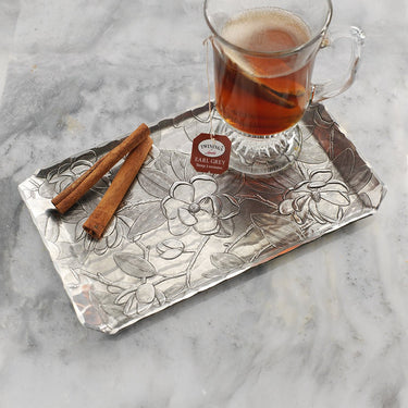 Clear glass cup of tea with cinnamon sticks on a decorative metal tray.
