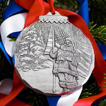 Decorative Patriotic Santa ornament with a winter scene and person holding an umbrella, hanging on red and blue ribbons.
