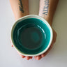 Teal ceramic bowl held by a person with tattoos on a light background