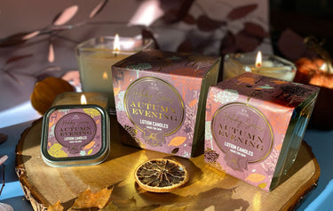 Two boxes of 'Autumn Evening' Soy Lotion Candle on a wooden surface with lit candles in the background.