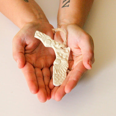 Two hands holding a white ceramic ornament shaped like a state on a plain background