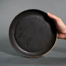 Hand holding a black ceramic plate against a gray background