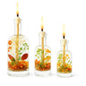 Three glass bottles with lit candles inside, containing floral and leaf decorations on a white background.